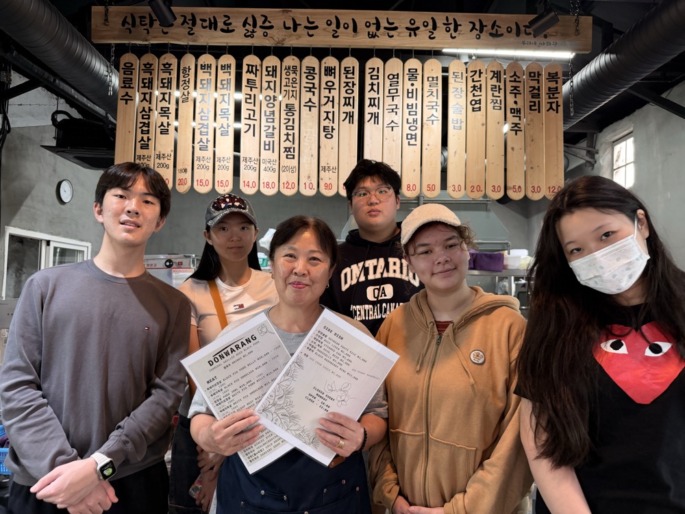 Owner of Donwarang holding the new meat and side-dish menus with students, under the wooden menu boards