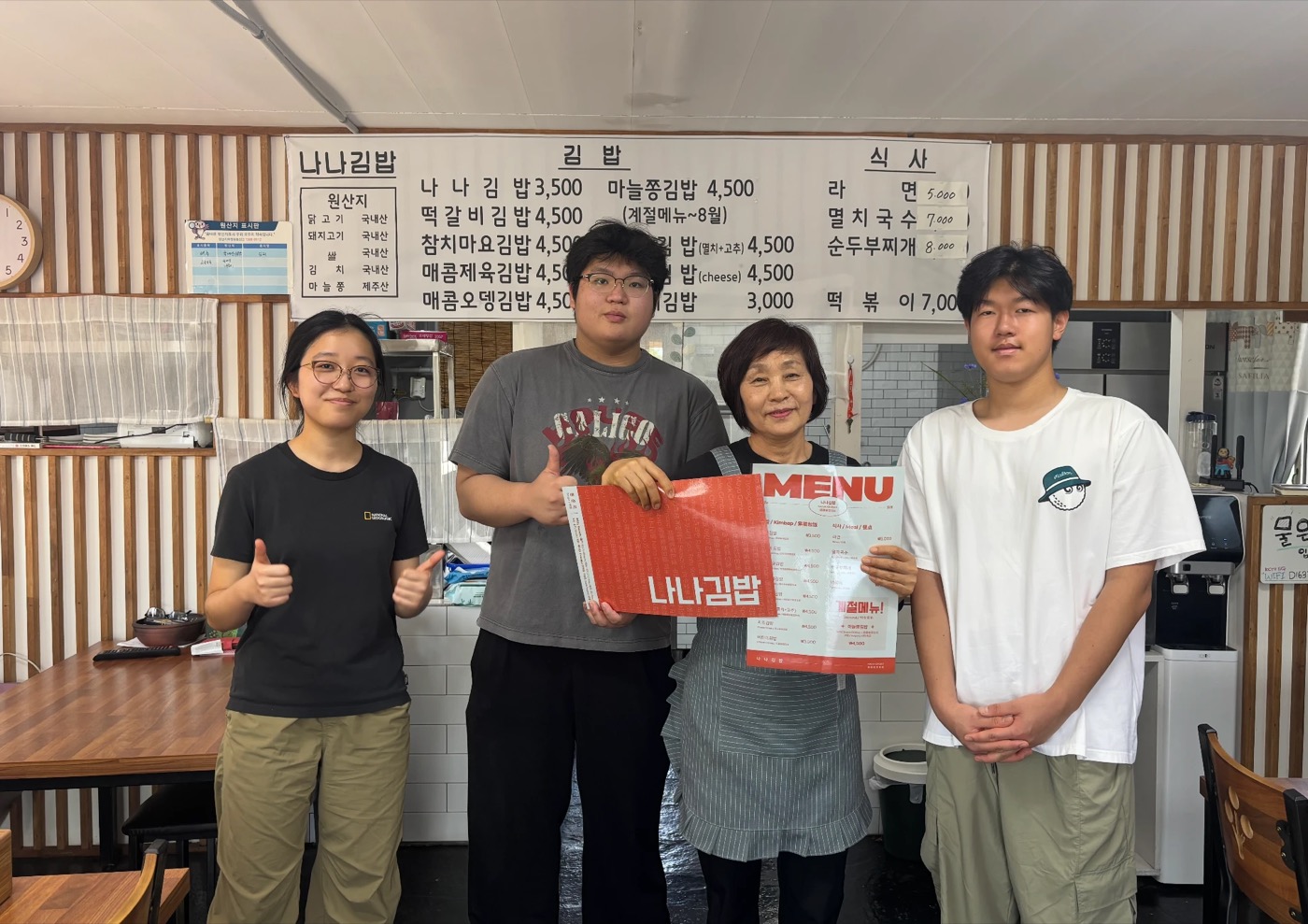Nana Gimbap's owner holding the new multilingual menu with three About Jeju students, the old handwritten menu board behind them
