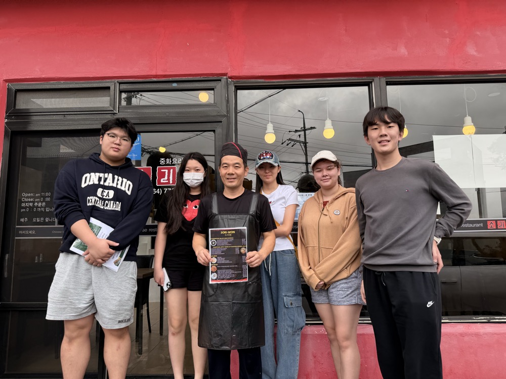Owner of Soriwon holding the new menu outside the red storefront with students